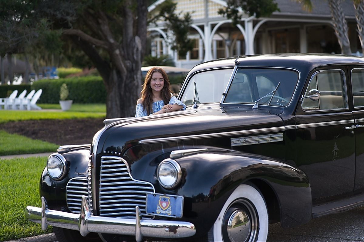 Senior portrait of a smiling teen girl posing with a classic vintage black 1940s Buick automobile outdoors.