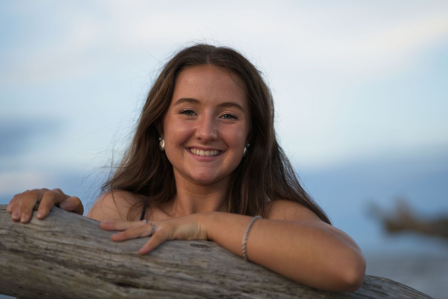 Senior portrait of a happy smiling teen girl with brown hair leaning on driftwood log at beach during sunset.