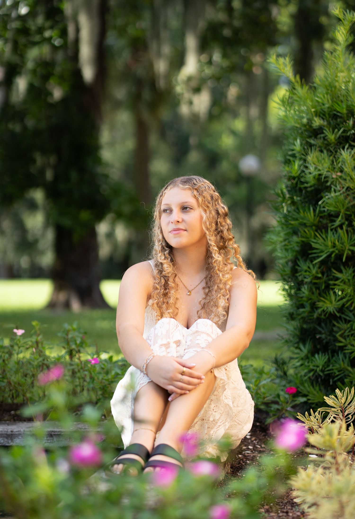 Young woman with curly hair in white dress sitting among pink flowers in a lush green garden.