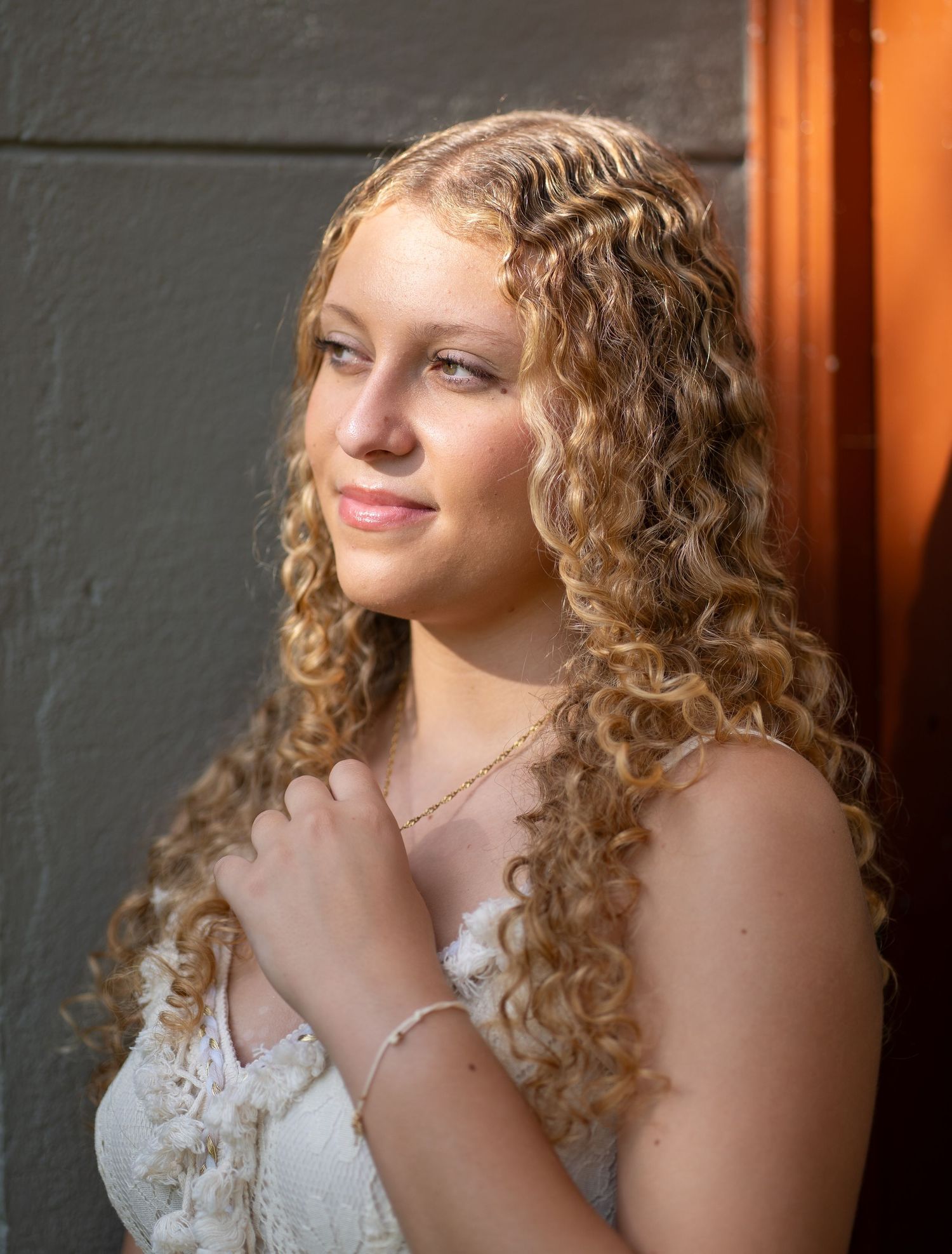 Young woman with long curly blonde hair wearing a white lace dress and bracelet, smiling softly outdoors.
