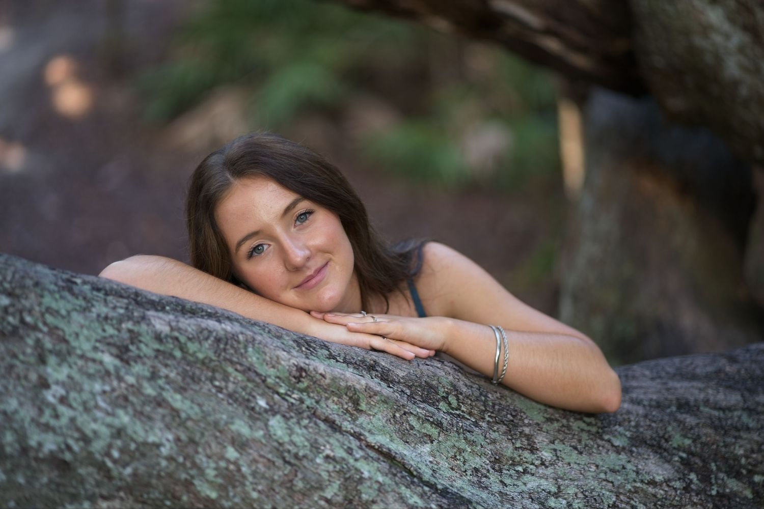 Senior portrait of a smiling young woman with brown hair resting chin on hands atop a large mossy rock.