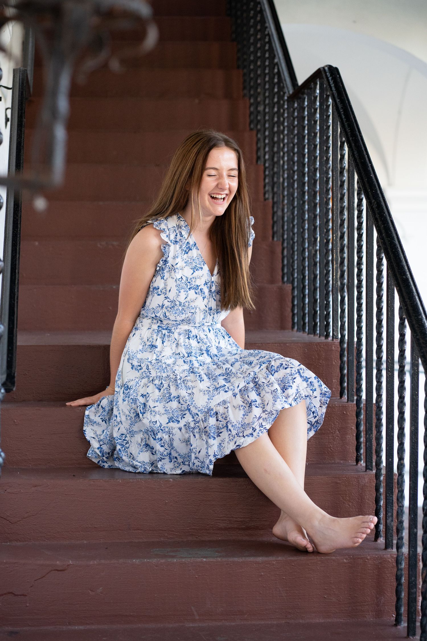 Young woman in blue floral dress laughing joyfully while sitting barefoot on outdoor brick stairs.