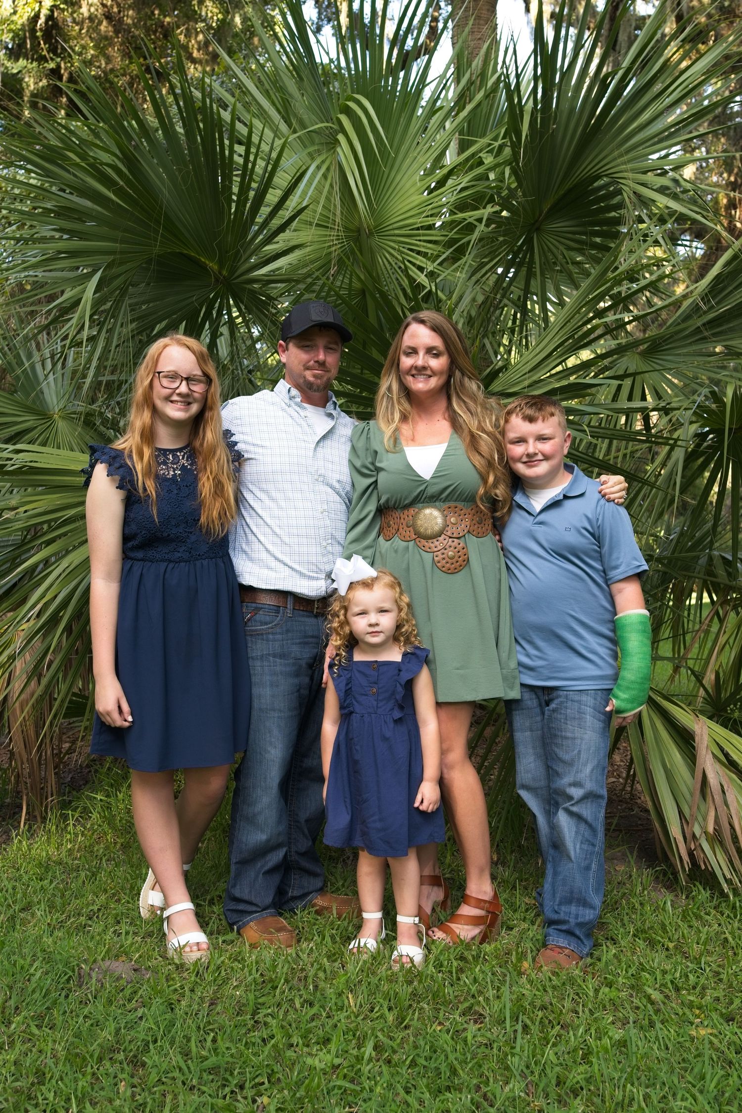 Family of five posing outdoors in coordinating blue and green outfits in front of tropical palm trees.
