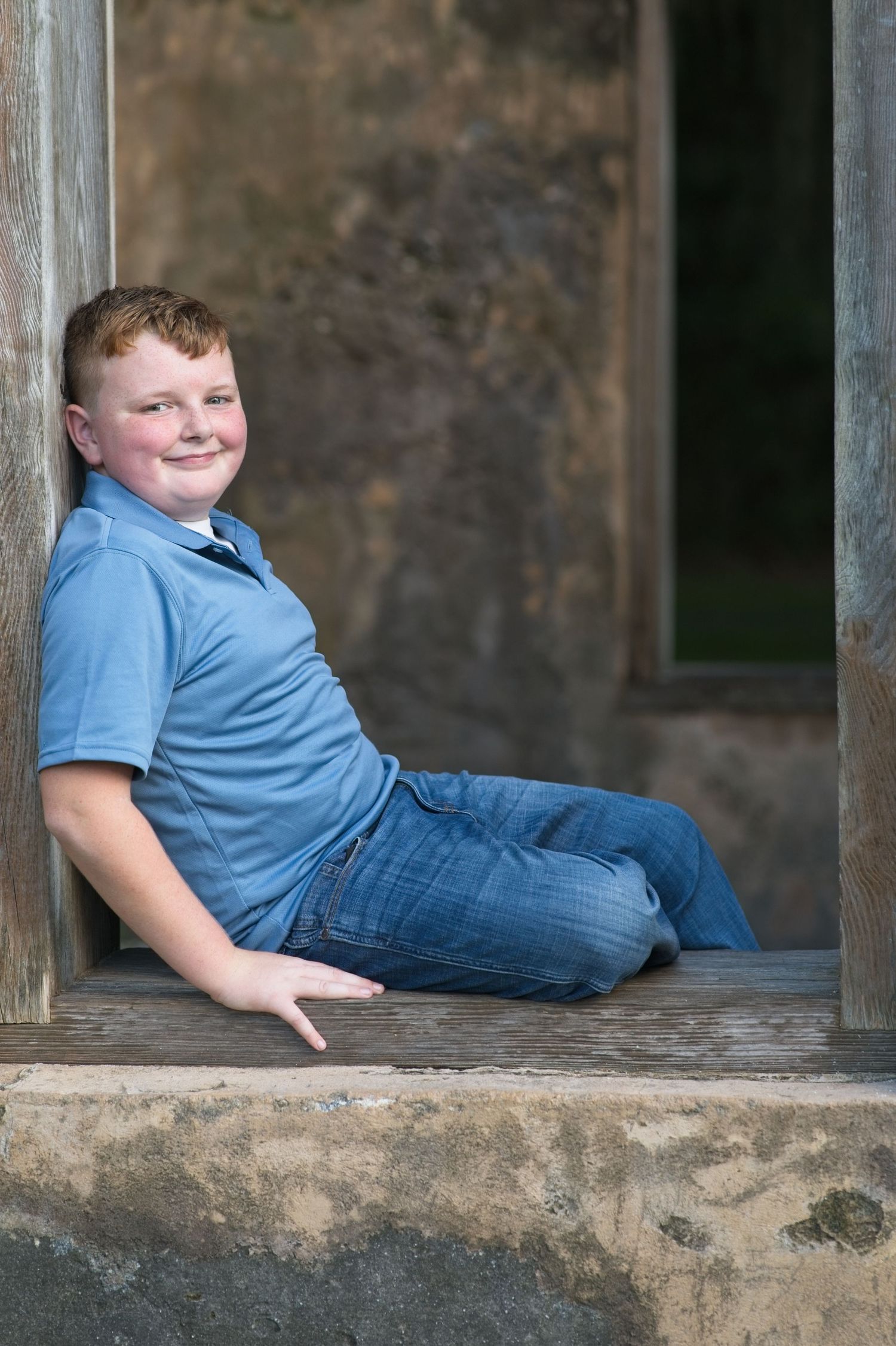 Smiling red-haired boy in blue polo shirt sitting relaxed in a rustic wooden doorway.