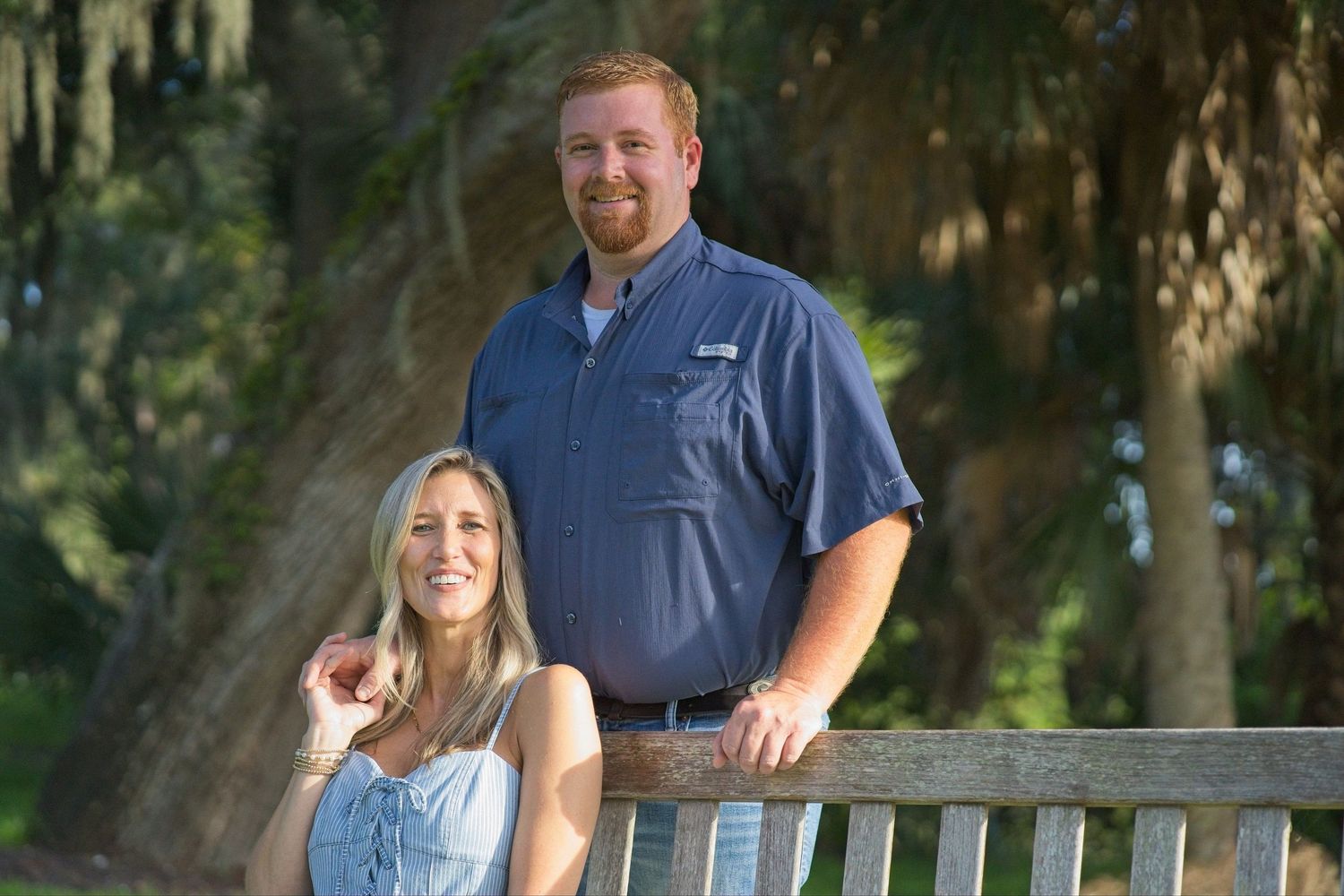 Smiling couple posing together outdoors near a wooden bench surrounded by lush green trees.