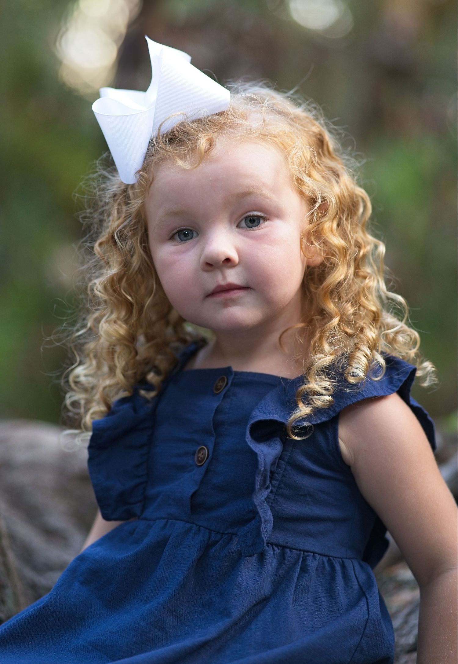 Adorable young girl with curly blonde hair wearing a navy dress and white bow hair accessory.