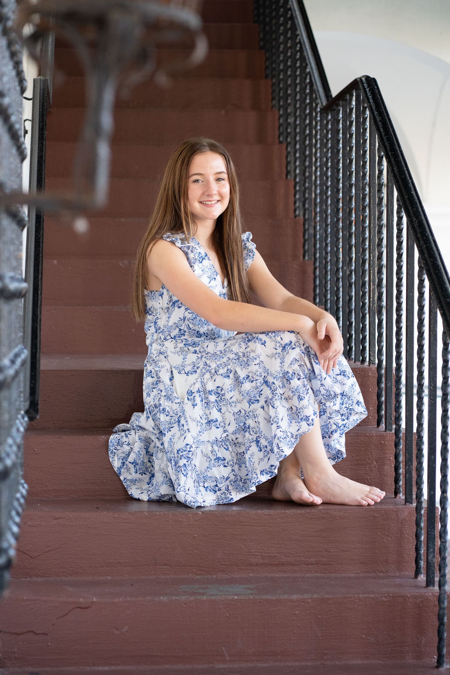 A smiling young girl in a blue floral dress sits barefoot on red brick stairs with black iron railings.
