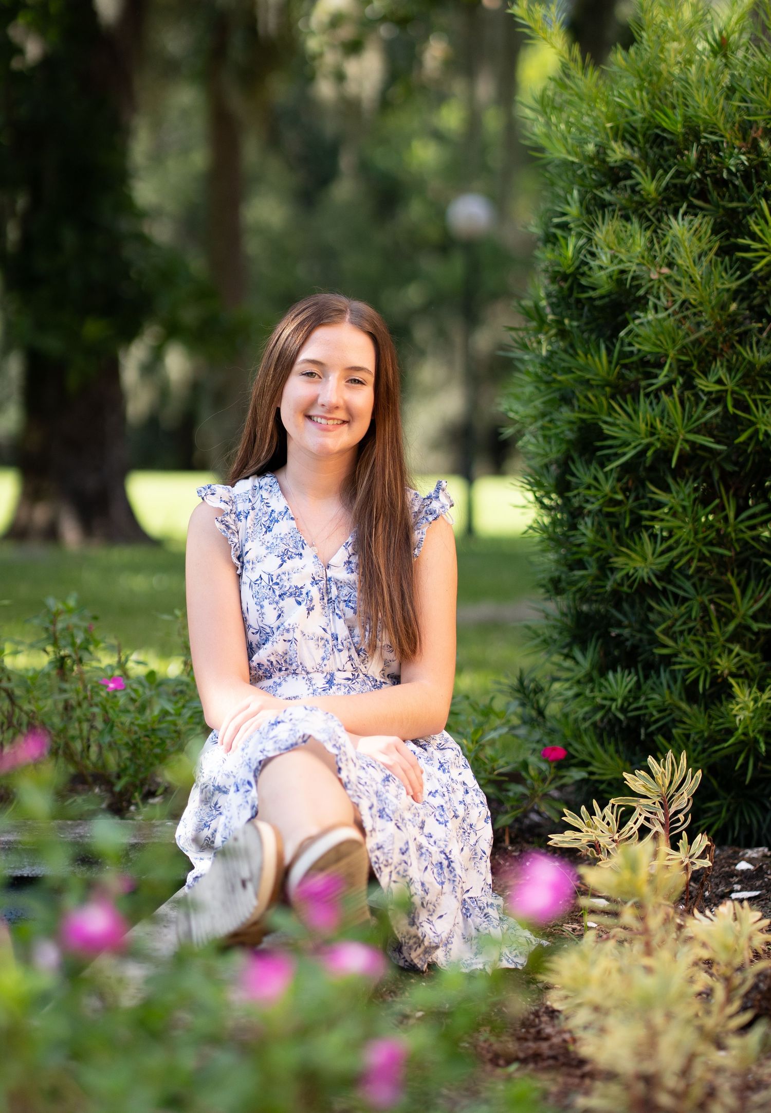 Young girl in floral dress sitting outdoors among pink flowers and lush greenery, smiling for a portrait photo.