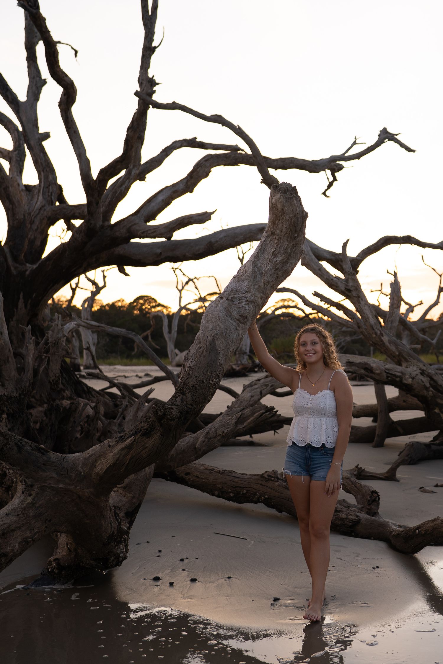 Young woman in denim shorts poses by driftwood trees on sandy beach at sunset.