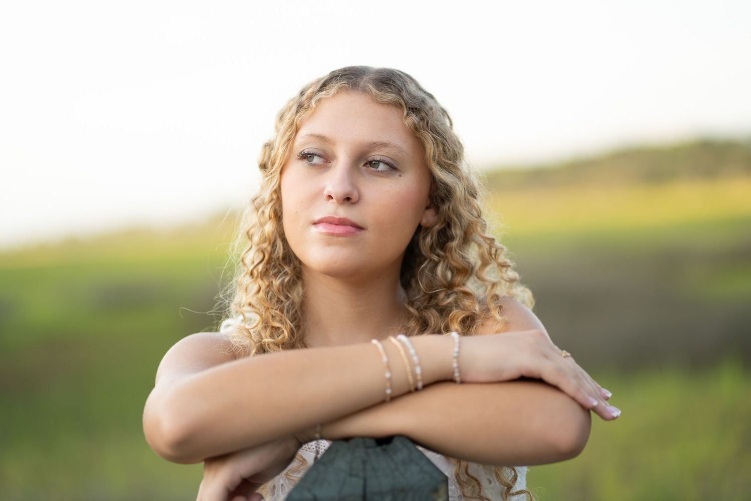 Young woman with curly blonde hair poses outdoors in a green field, arms crossed, wearing a denim top.