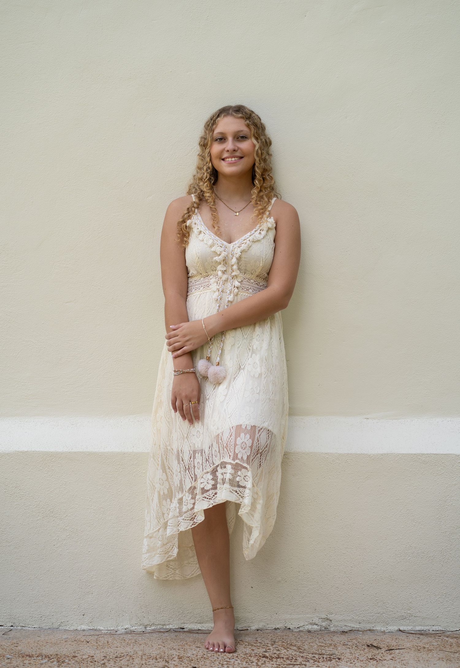 Young woman in cream lace midi dress smiling barefoot against a pale green wall.