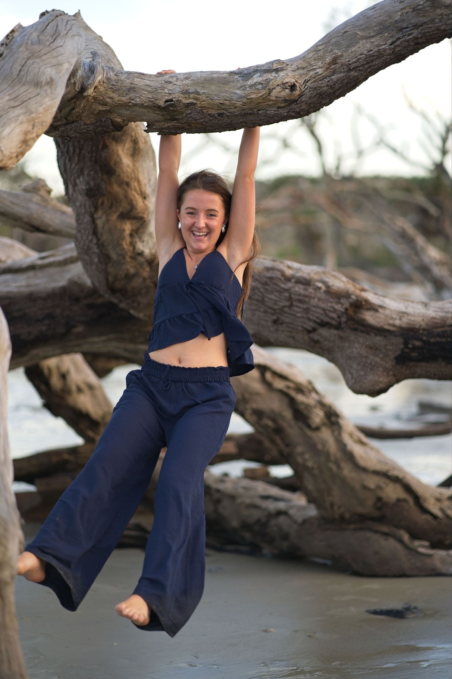 Senior portrait of a laughing young woman hanging from driftwood branches at beach wearing navy crop top and wide-leg pants.