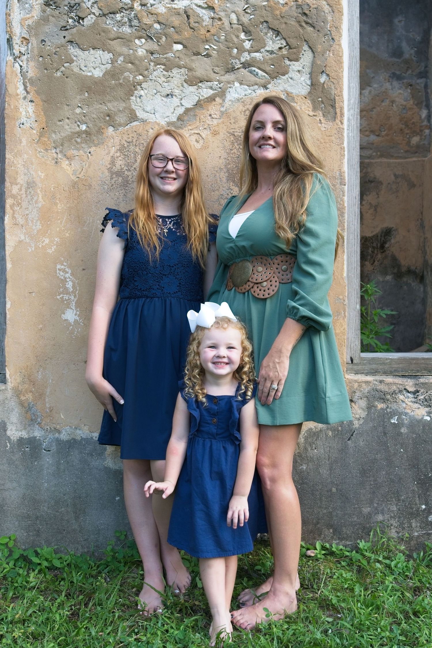 Mother and two daughters in matching navy blue dresses posing against a rustic weathered wall.
