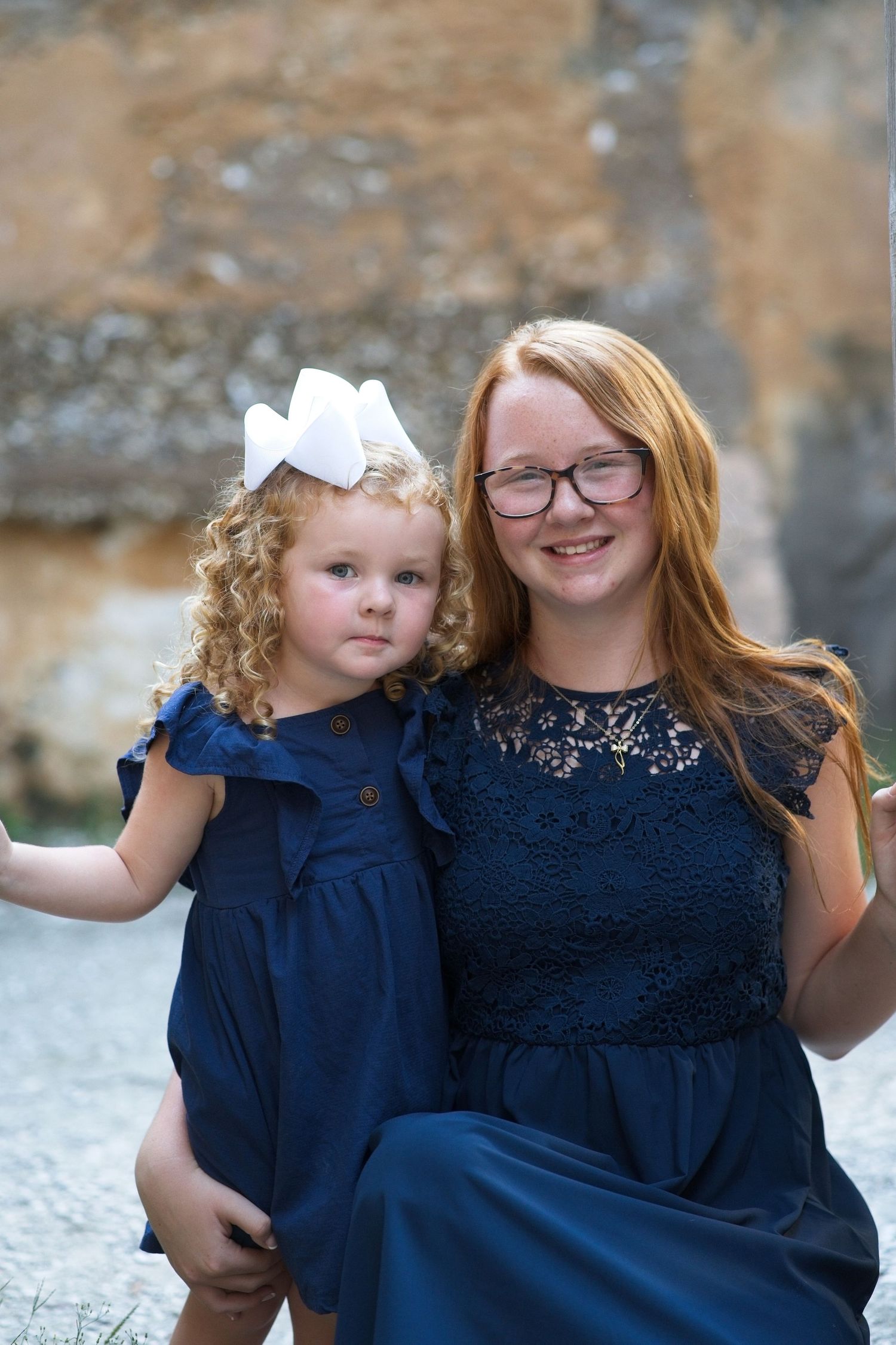 Teenage girl with red hair and glasses hugging her curly-haired little sister in navy blue dresses.
