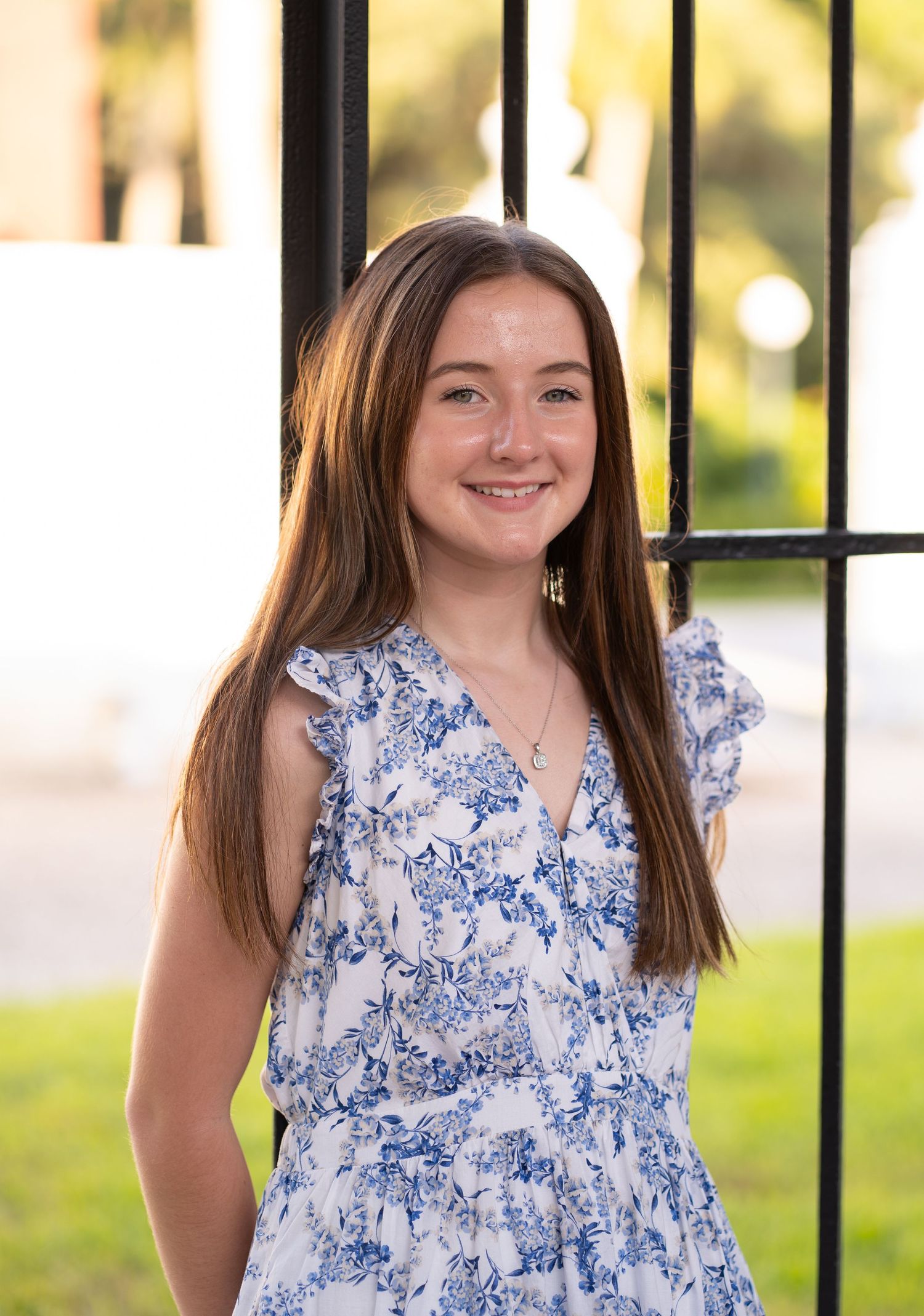 Smiling teenage girl in blue floral dress leaning against black iron gate outdoors.