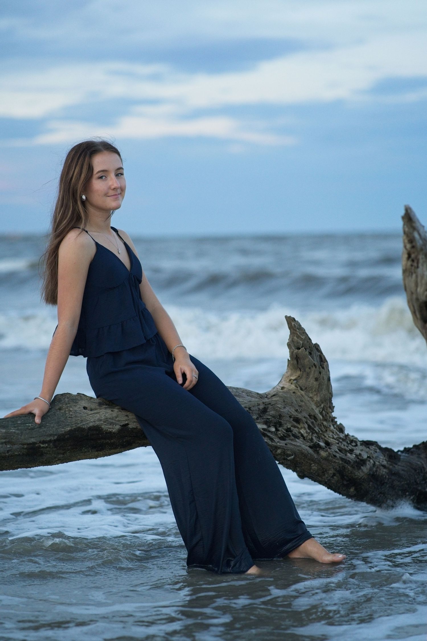 Senior portrait of a teen girl in navy jumpsuit sitting on driftwood at the beach with ocean waves behind her.