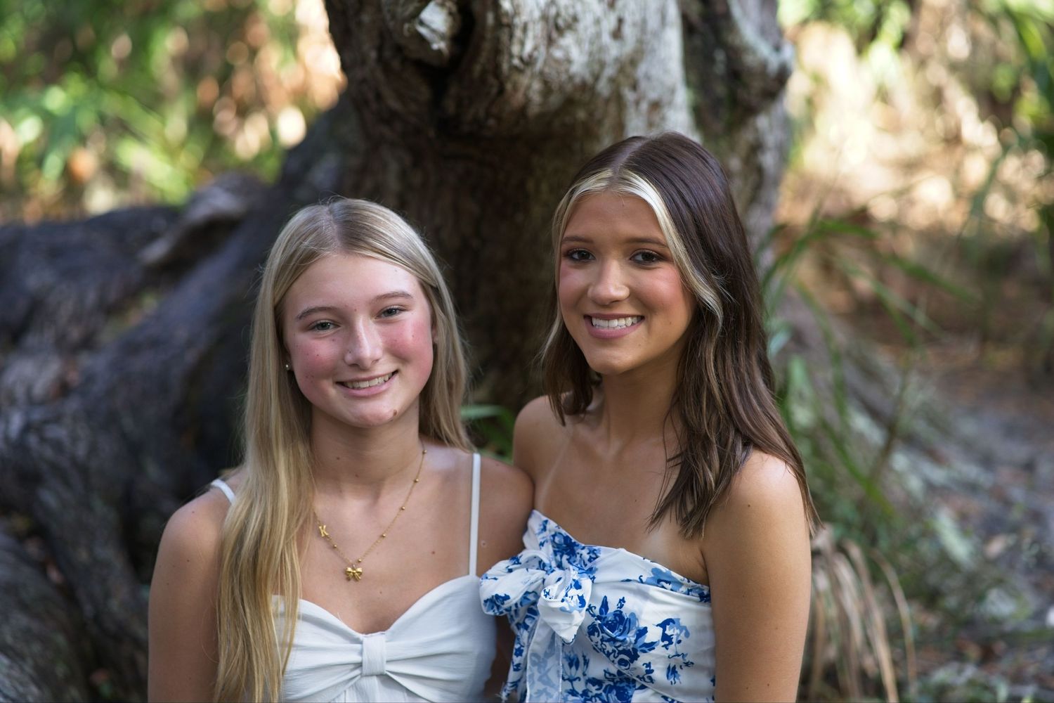 Two smiling teenage girls posing together outdoors in front of a large tree in summer dresses.