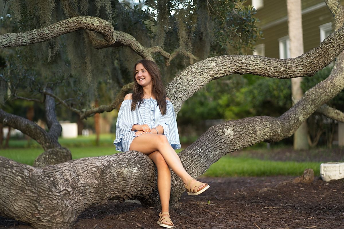 Senior portrait of a young woman in blue off-shoulder dress sitting on large twisted oak tree in park setting.