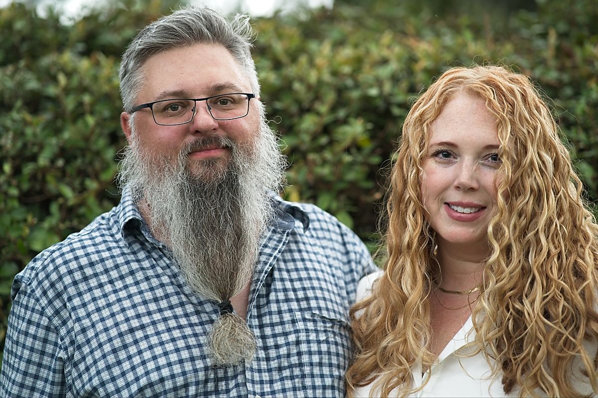 Close-up portrait of a bearded man and curly-haired woman smiling together outdoors near green hedges.