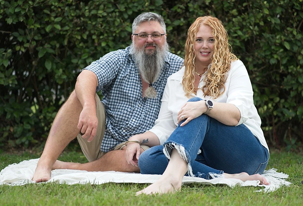 Couple sitting casually on a white blanket in green grass, smiling together outdoors.