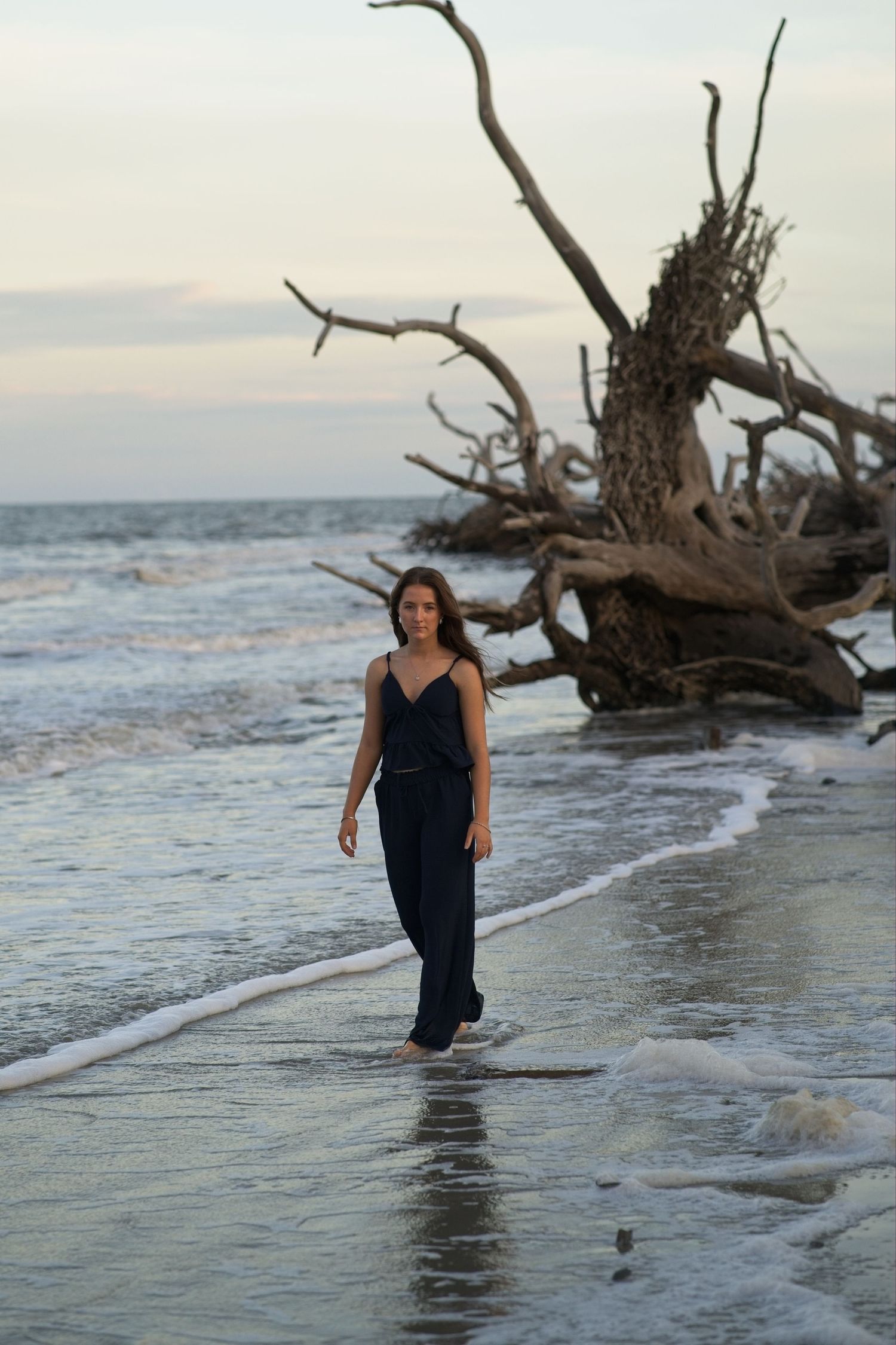 Senior portrait of a young woman in navy jumpsuit walking barefoot along shoreline with dramatic driftwood behind her.