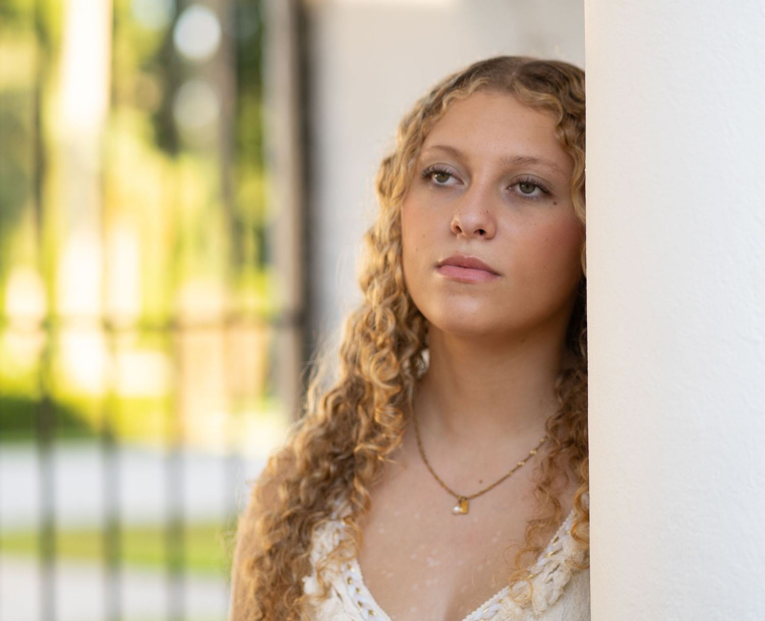 Young woman with curly blonde hair leans against a white pillar, wearing a delicate necklace outdoors.