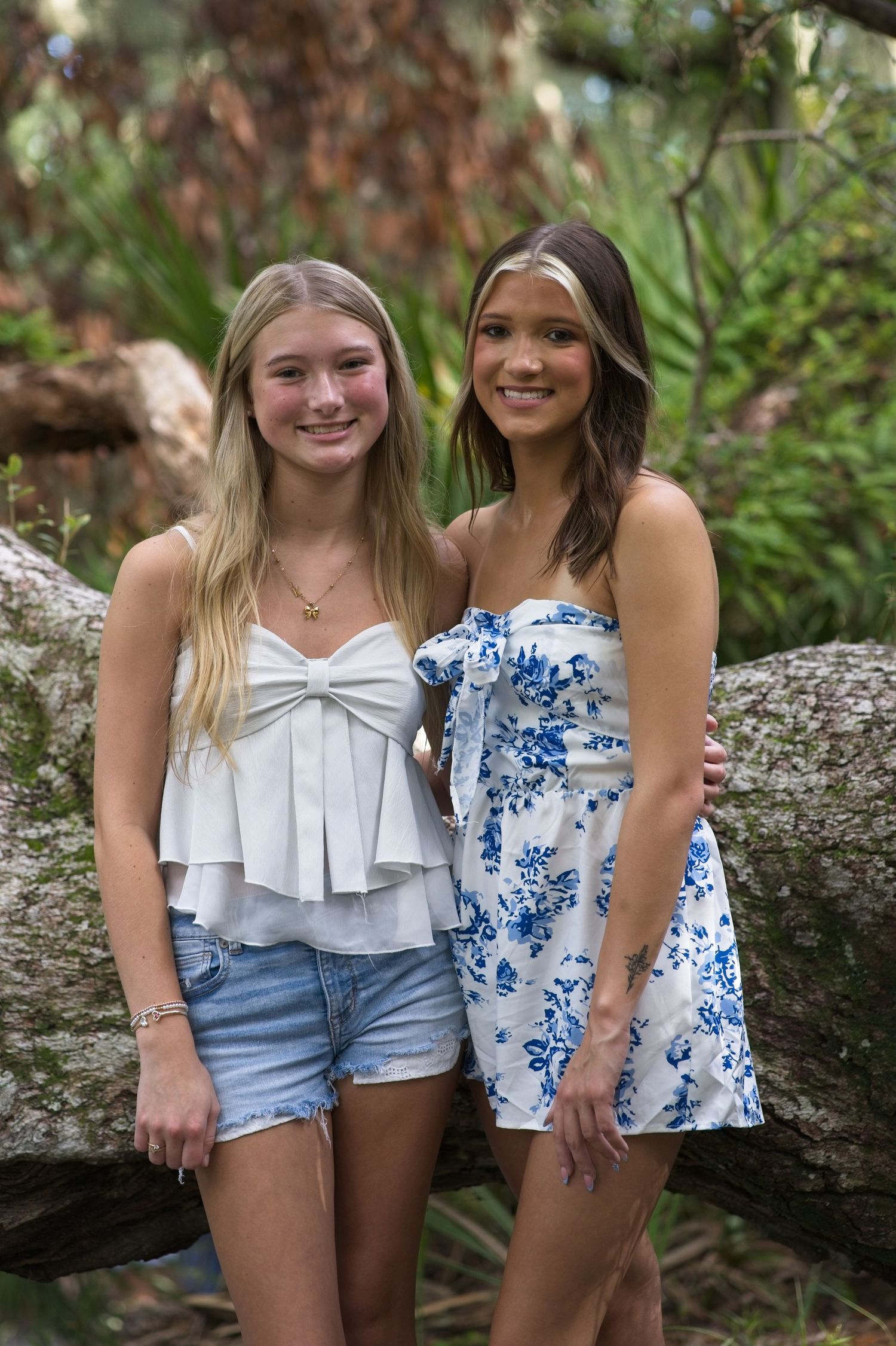 Two teenage girls smiling together outdoors, one in white top with denim shorts, other in blue floral dress.