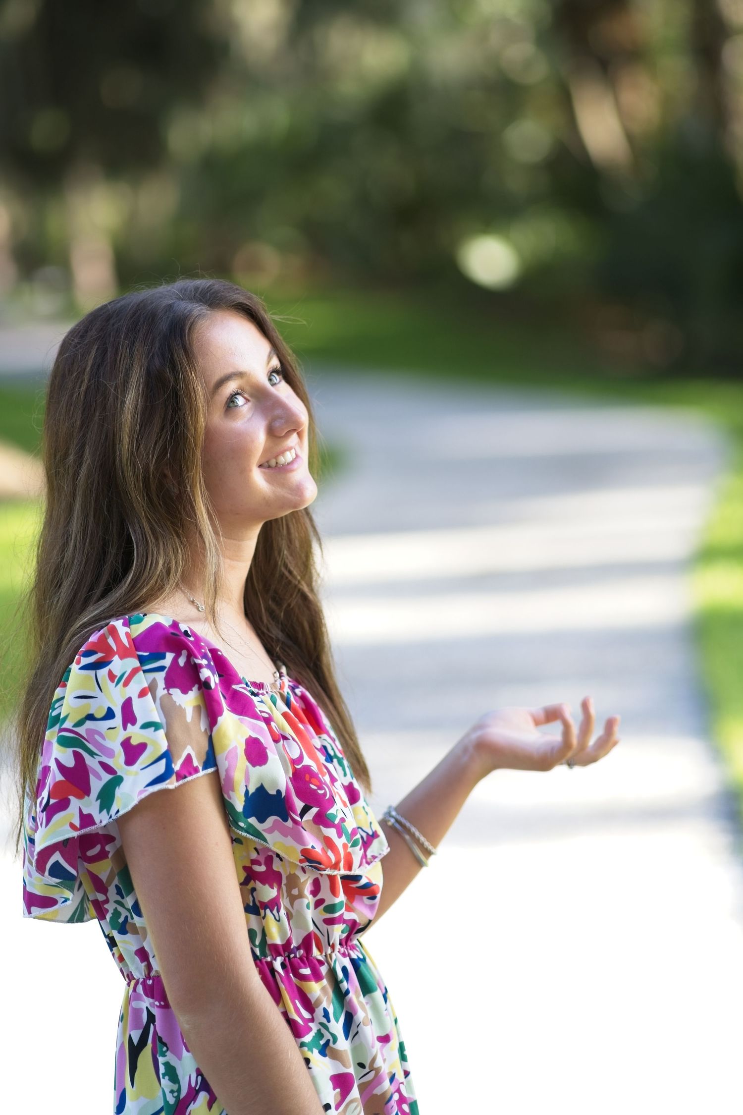 Senior portrait of a happy young woman in colorful floral dress smiling upward with hand extended in sunny park.