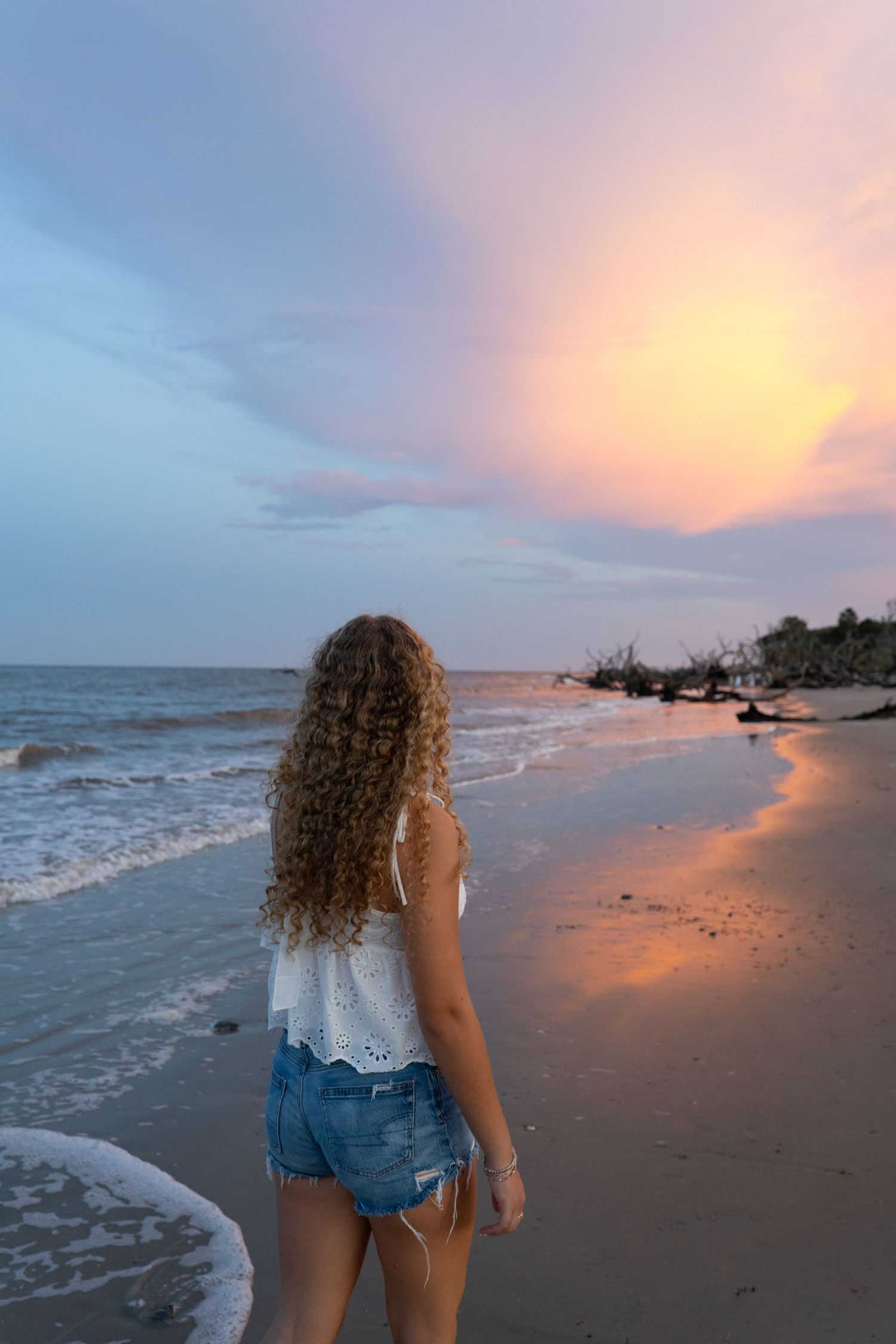 Young woman with curly hair in white top and denim shorts watching a colorful sunset on a sandy beach.