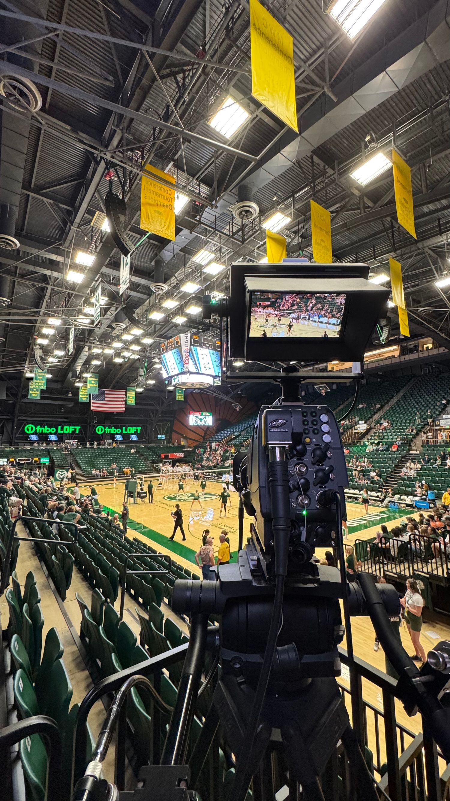 Professional video camera set up to film a college basketball game in a green arena.