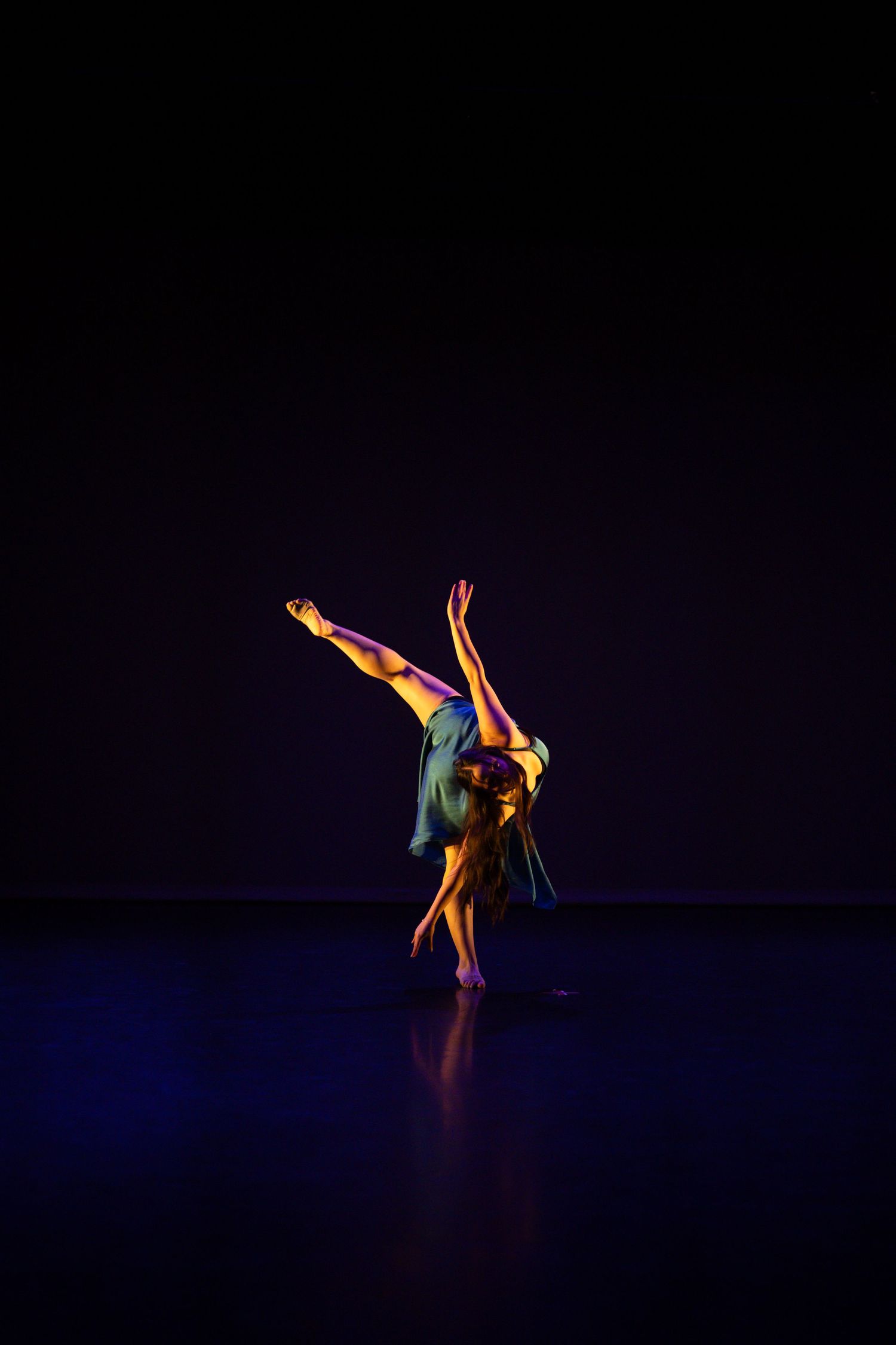 Solo contemporary dancer performs an elegant backbend with leg extended under dramatic stage lighting.