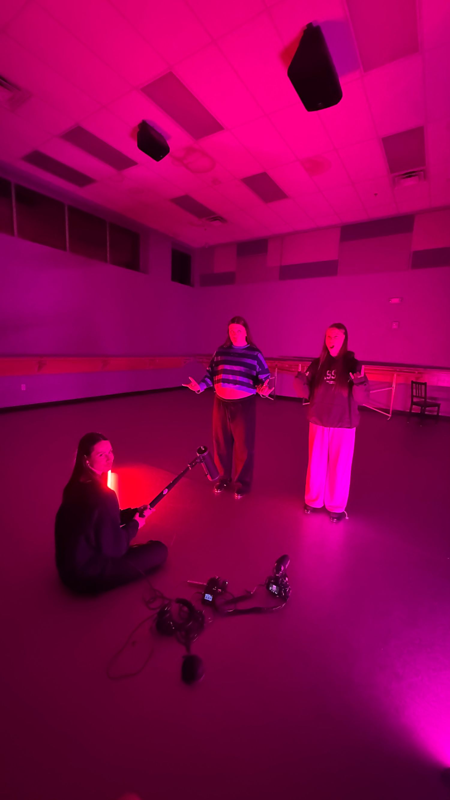 Three students filming a video production in a pink-lit empty dance studio.