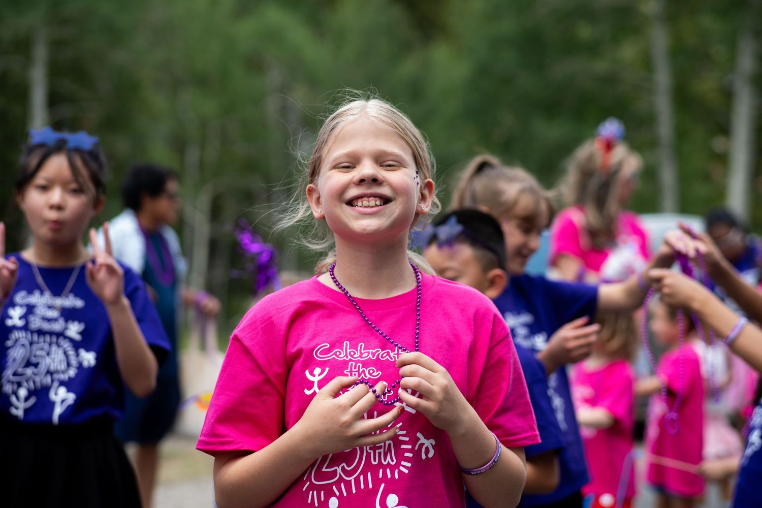 A joyful blonde girl in a pink camp t-shirt smiles outdoors at a summer camp event.
