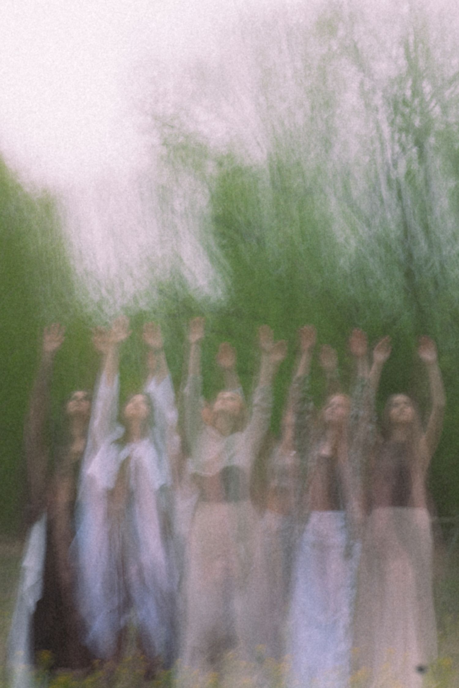 A blurred long-exposure photo captures multiple dancers raising their arms in a lush green outdoor setting.