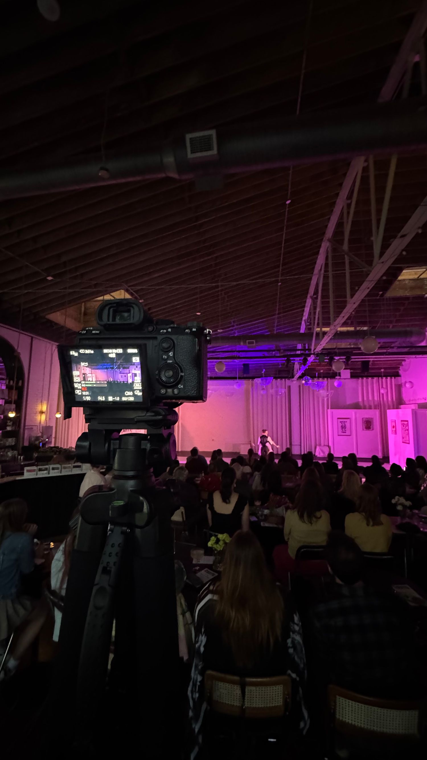 Camera on tripod filming a live indoor event with purple stage lighting and seated audience.