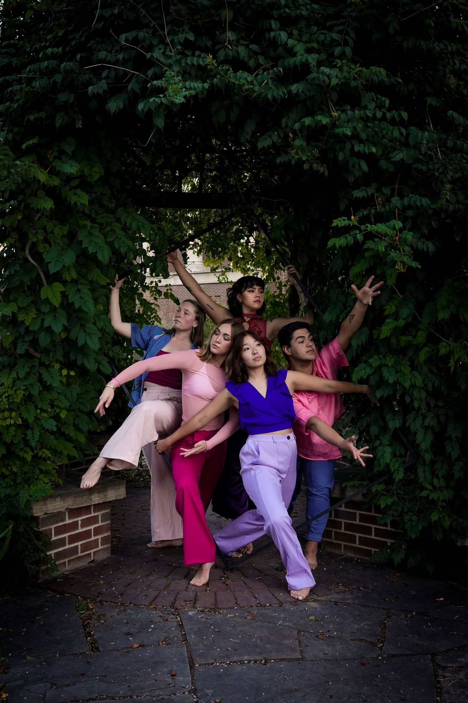 Five dancers pose gracefully outdoors against a lush green ivy-covered wall in vibrant clothing.