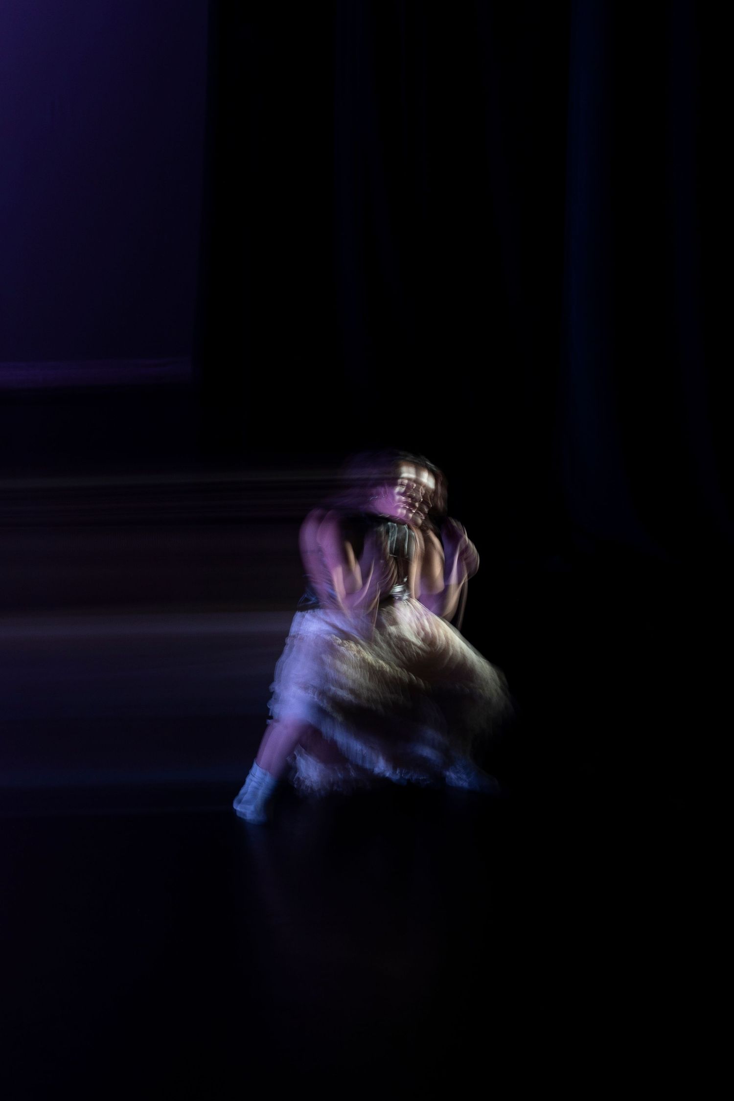 A solo dancer in purple and white costume spins in a long-exposure motion blur photograph on a dark stage.