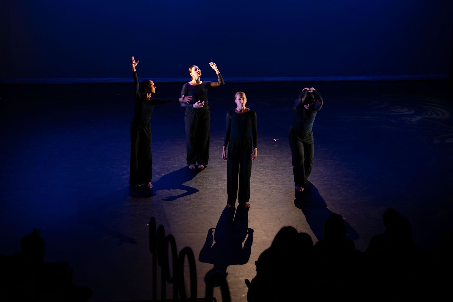 Three dancers in black perform on a dimly lit stage with blue lighting, arms raised expressively.