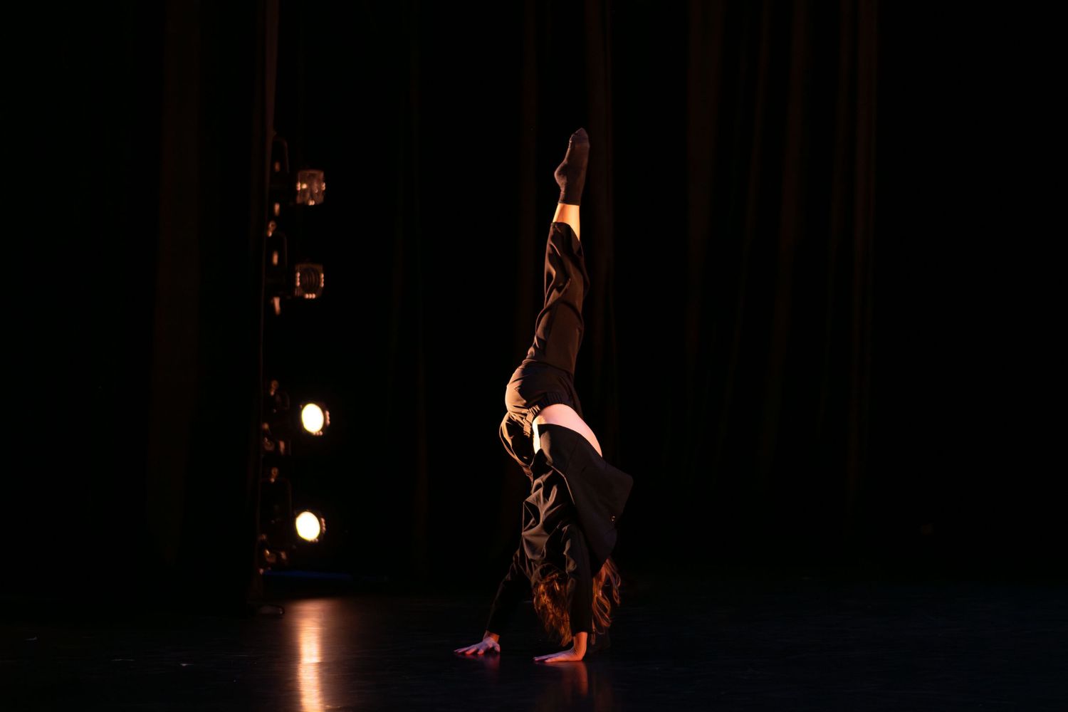 Dancer performs a headstand silhouetted against stage lights in a dark theater.