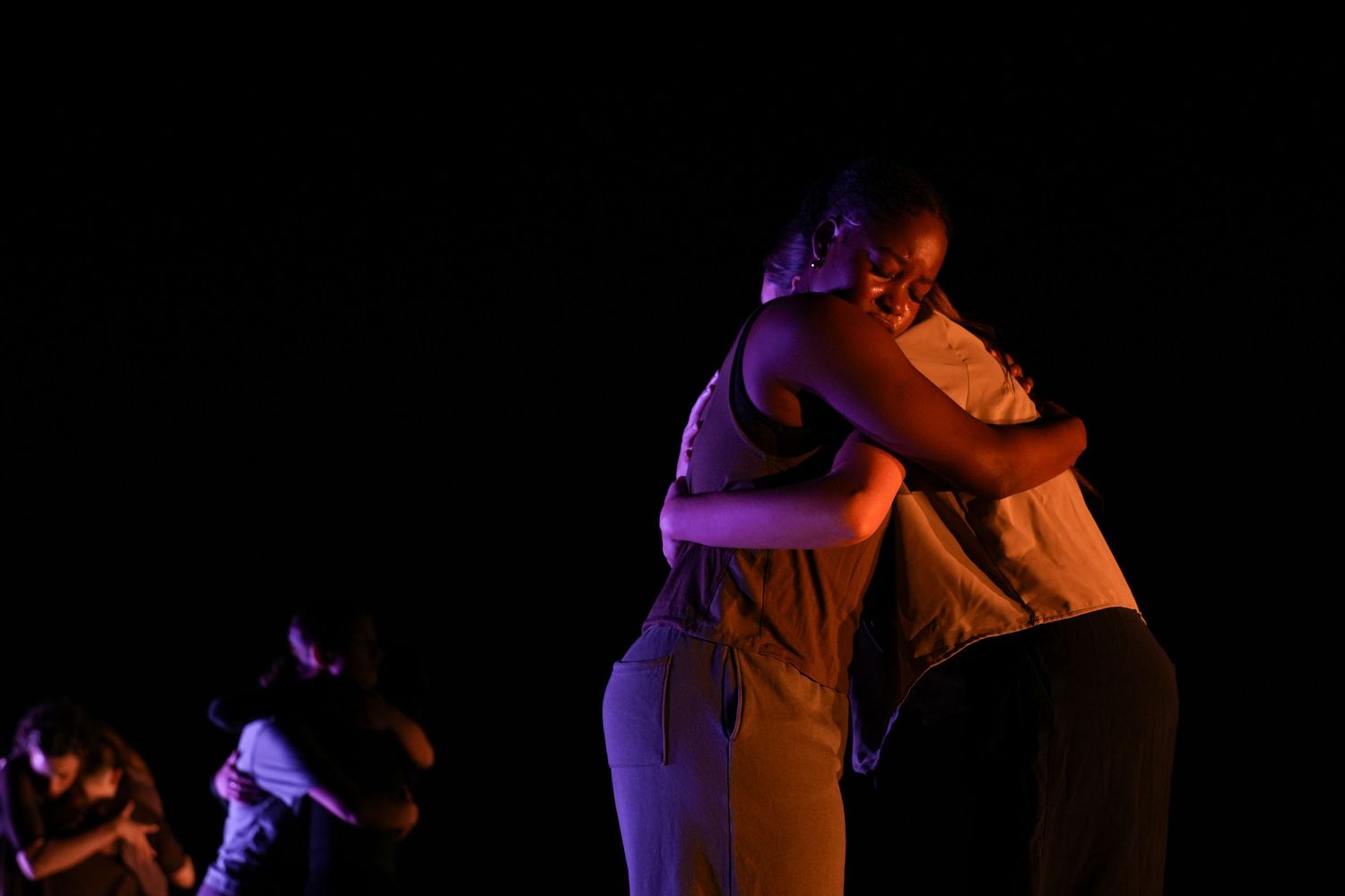 Two dancers share an emotional embrace on a darkened stage illuminated by warm purple and blue stage lighting.
