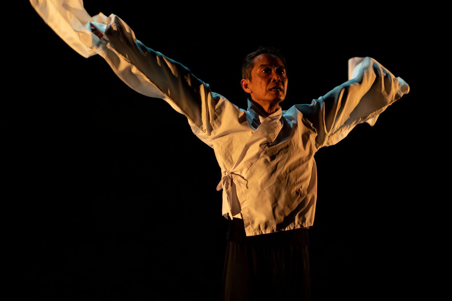 A male dancer spreads arms wide in a white shirt, captured mid-performance against a black backdrop.