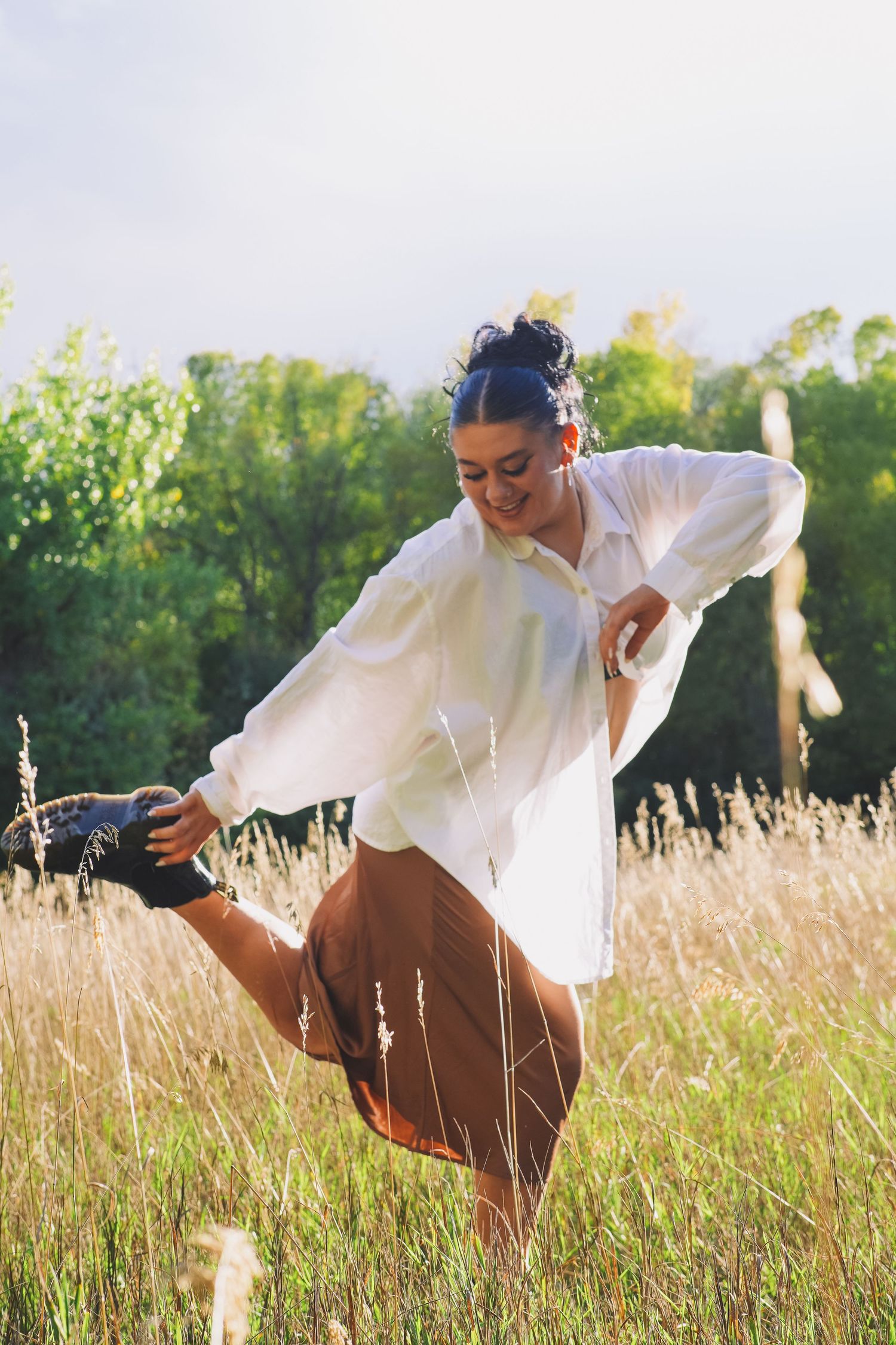 A smiling dancer in a white shirt and brown skirt performs a high kick in a sunlit grassy field.
