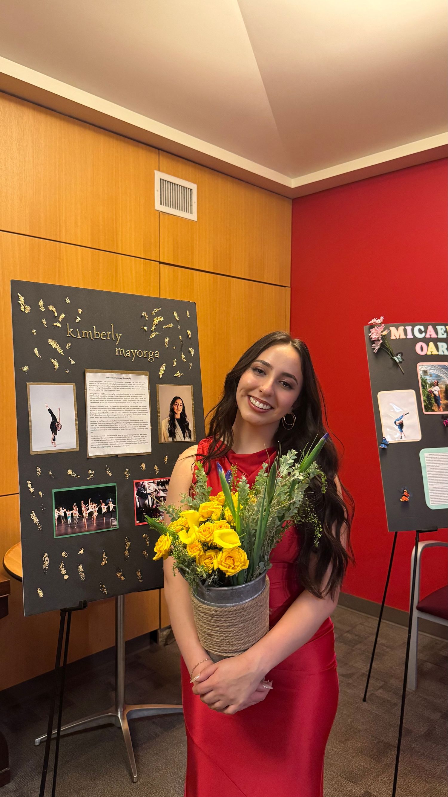 Kimberly Mayorga in red dress holding yellow flowers stands before her dance showcase display board.