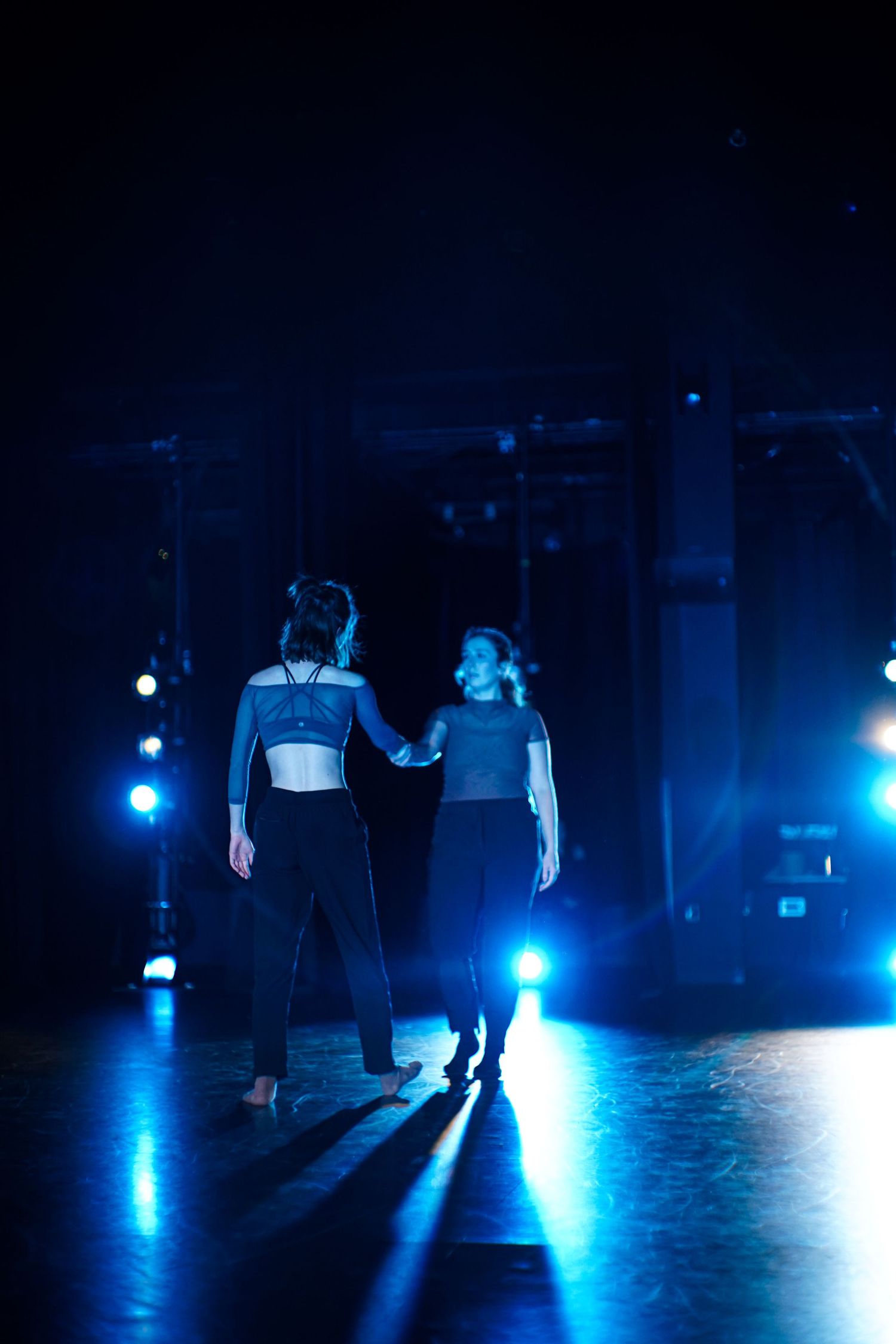 Two female dancers hold hands on a dimly lit stage with dramatic blue lighting during a contemporary dance performance.