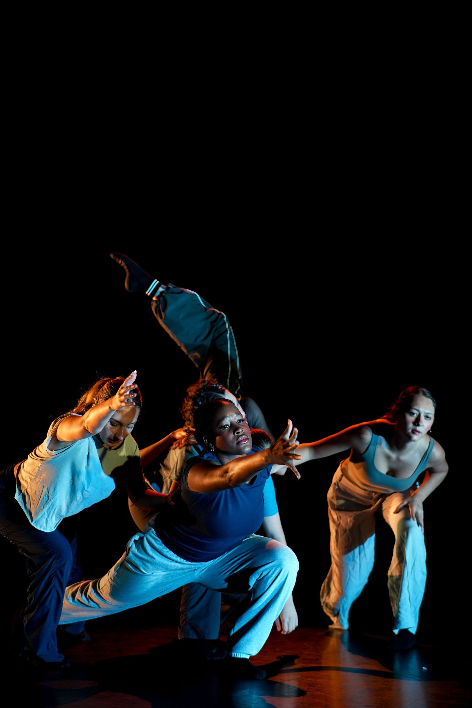 Three dancers perform an expressive contemporary routine under colorful stage lighting on a dark stage.