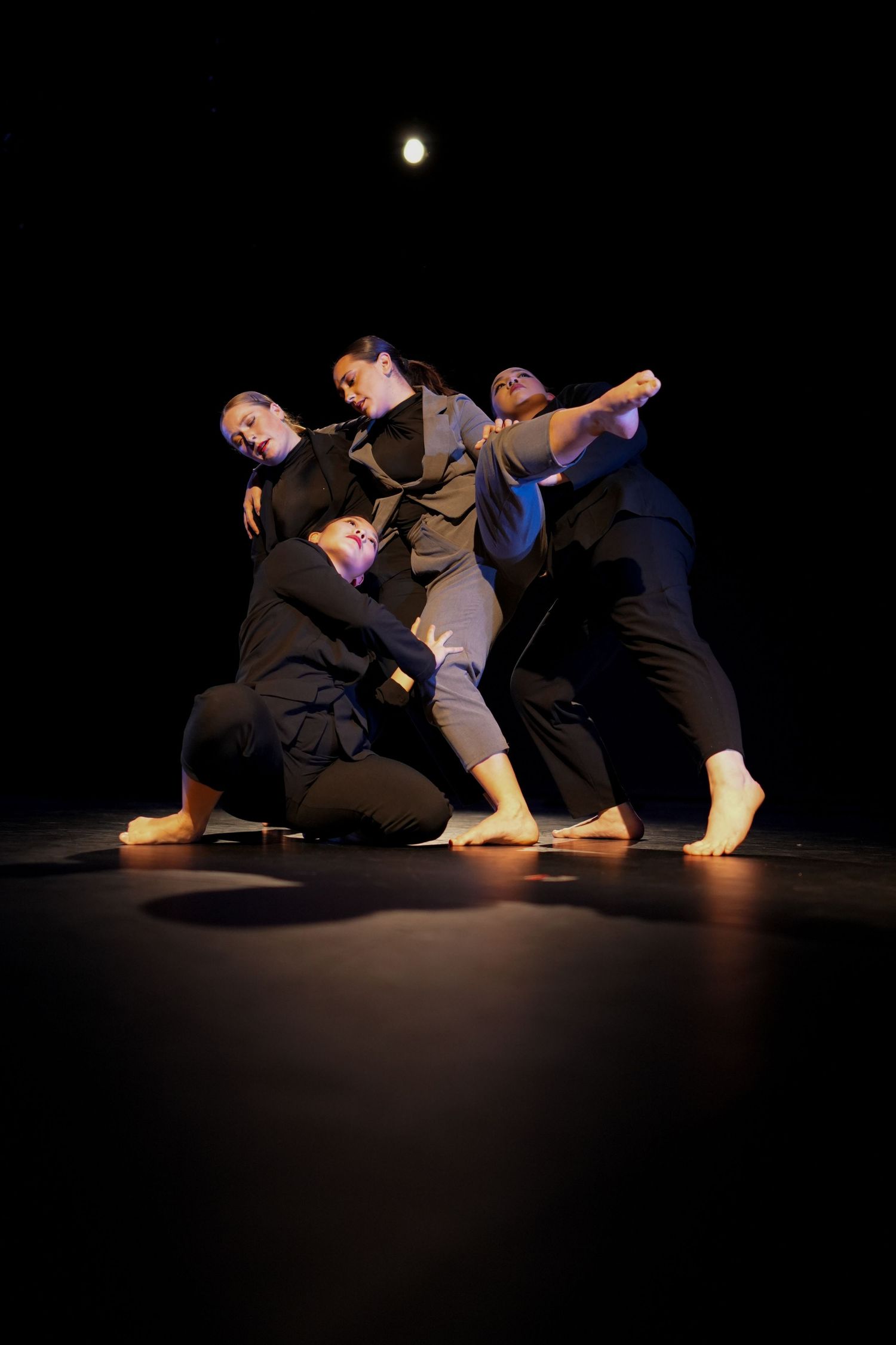 Four contemporary dancers form a close group pose on a black stage under warm lighting.