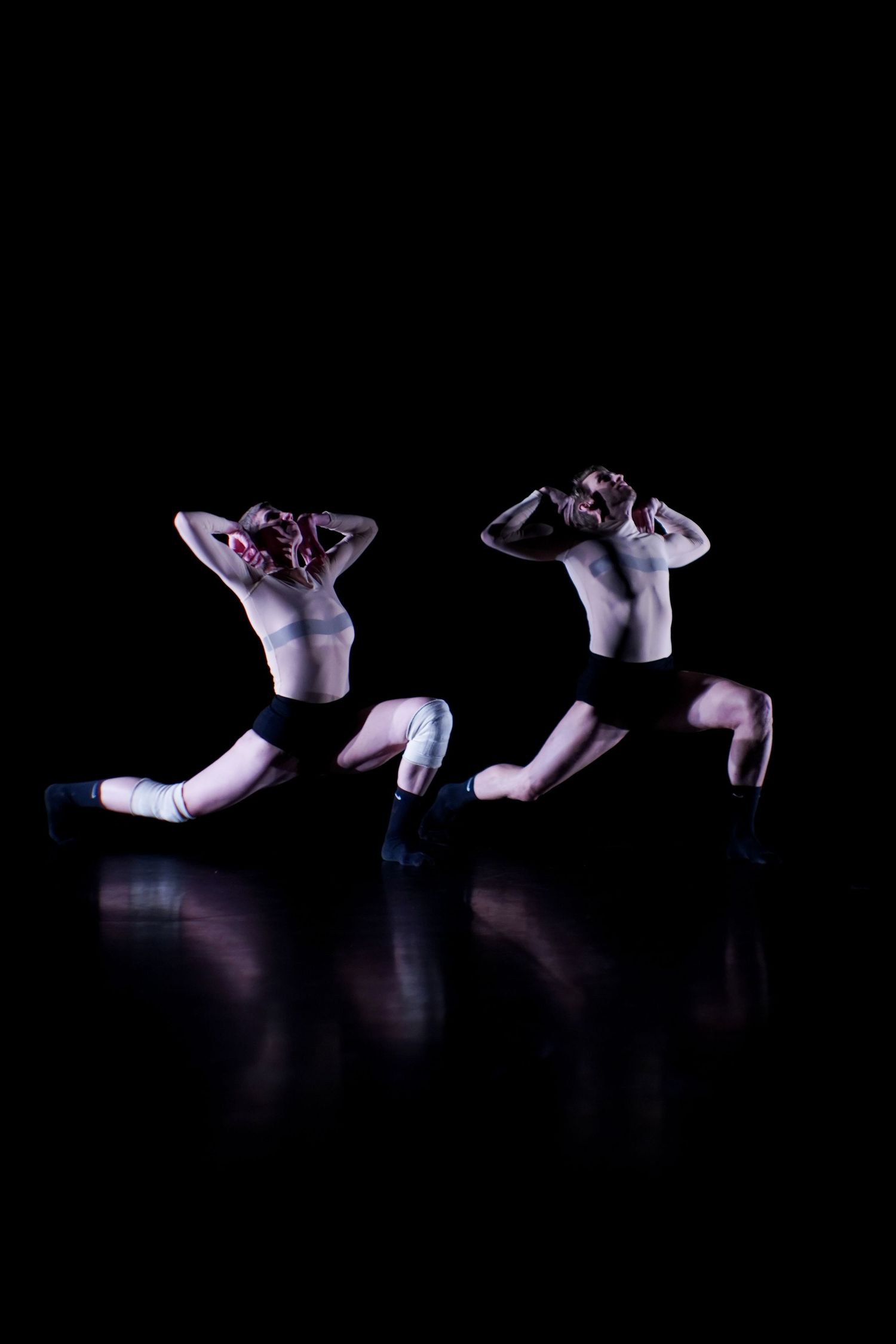 Two synchronized dancers in black shorts sit mirrored on a reflective dark stage with dramatic lighting.