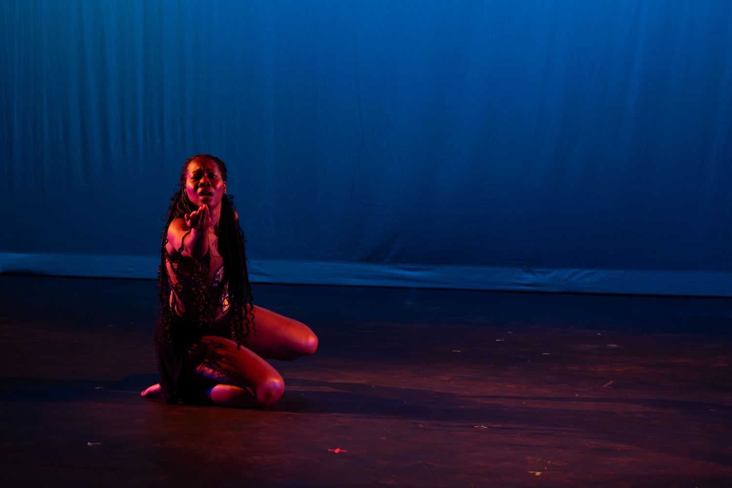 Female dancer sits on stage floor in an emotional pose under dramatic red and blue lighting.