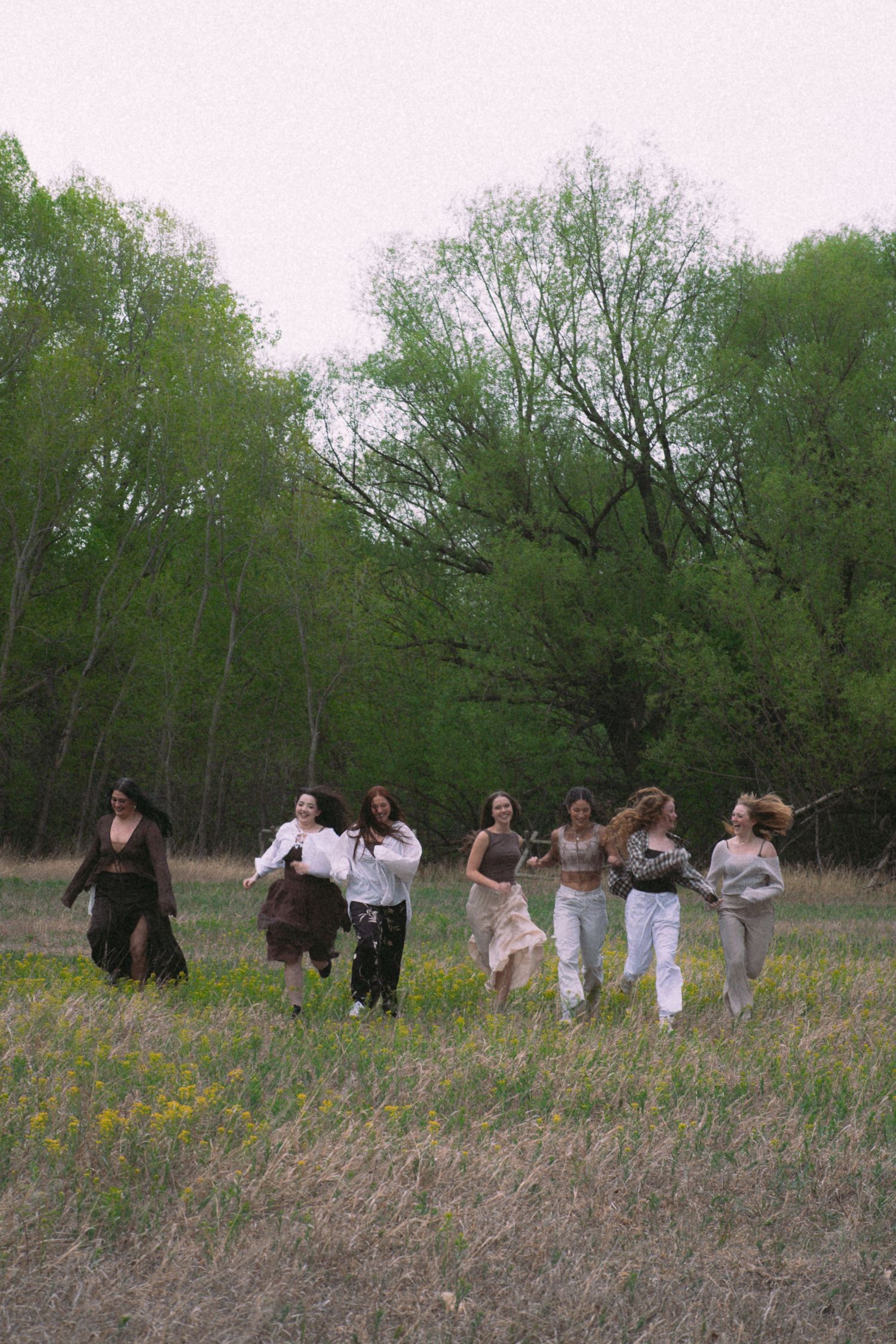 A group of seven women run freely through a lush green meadow surrounded by spring trees.