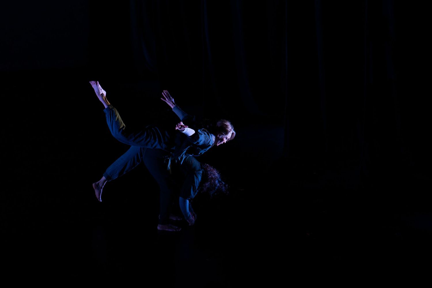 Two dancers perform a dramatic contemporary dance duet under blue stage lighting against a dark background.